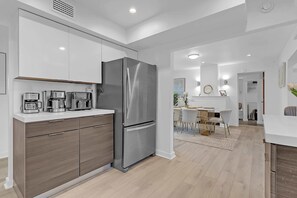 Een moderne keuken met een roestvrijstalen koelkast, een ingebouwde oven en een magnetron. Er is een houten kast met lades en een wit aanrecht. De keuken loopt over in een eethoek met een witte tafel en stoelen.