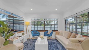 Un salón moderno con un sofá blanco, sillones azules, una mesa de centro blanca y ventanas amplias con vista a palmeras.