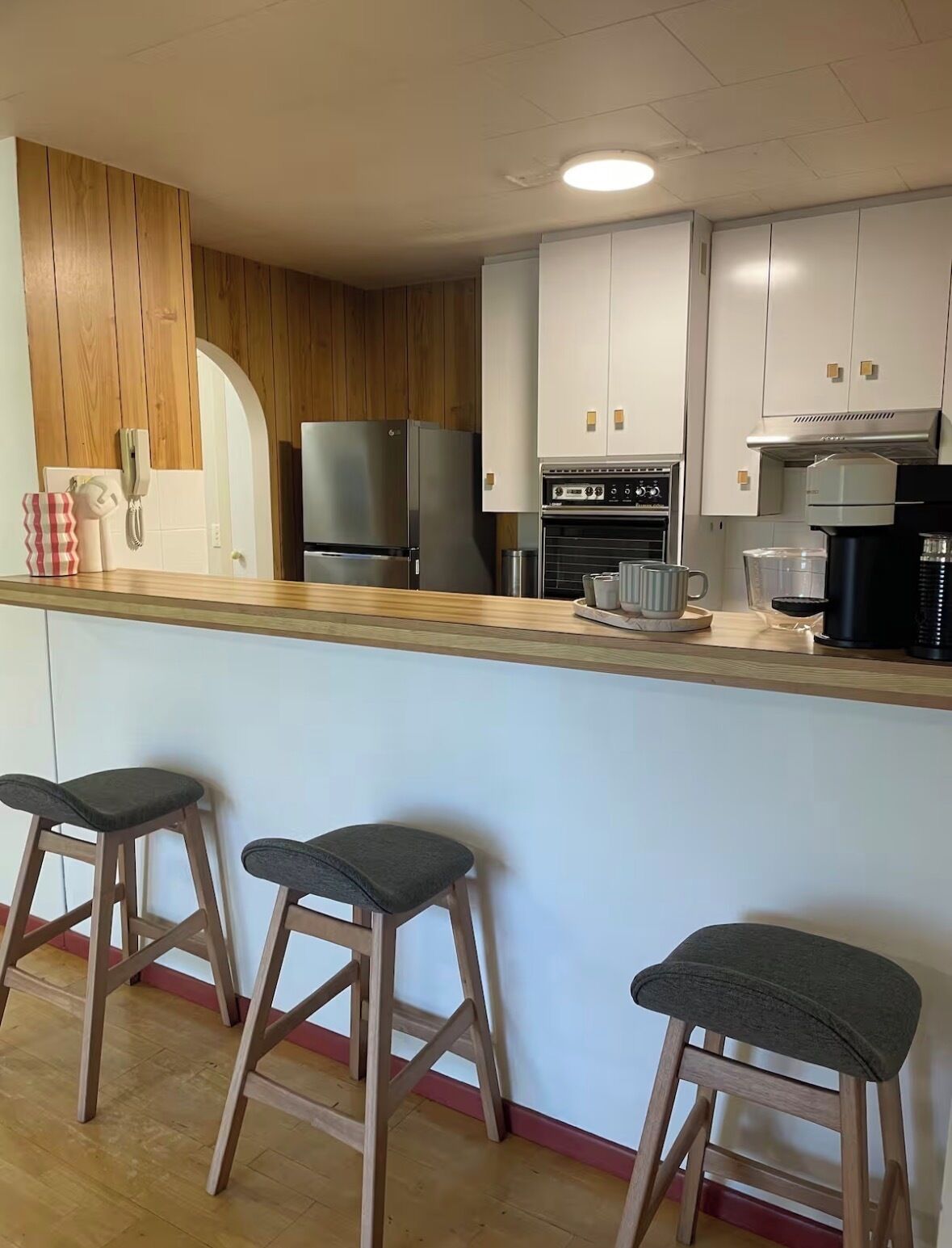 A modern kitchen with wooden accents, a built-in refrigerator, an oven, and a coffee maker. There are three bar stools and a countertop with various kitchen items.