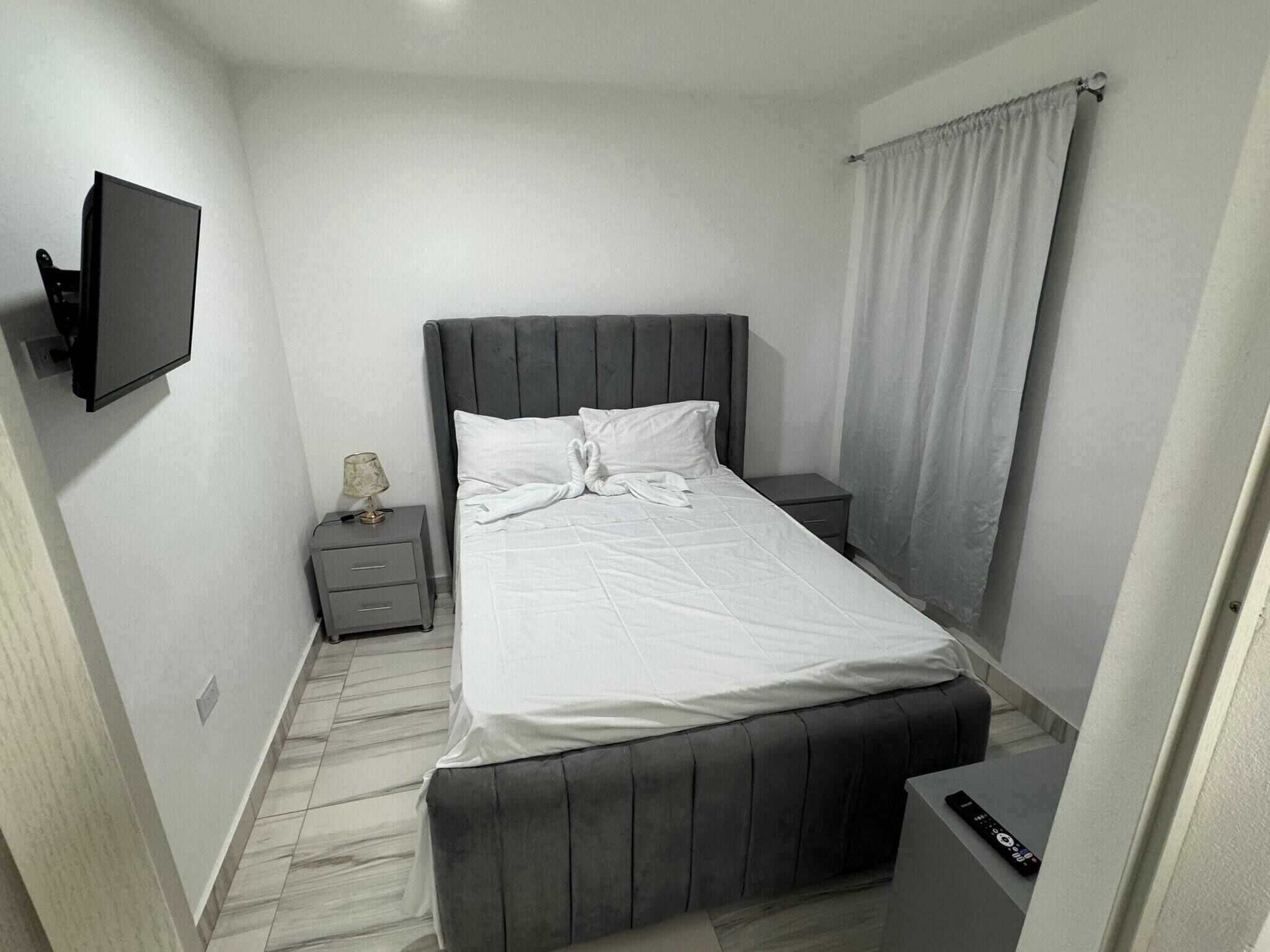 A bedroom with a bed, a bedside table, a lamp, a TV, and a curtain.
