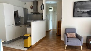 Een moderne keuken met witte kasten, een roestvrijstalen afzuigkap en een magnetron. In de woonruimte staat een gele prullenbak en een grijze fauteuil.