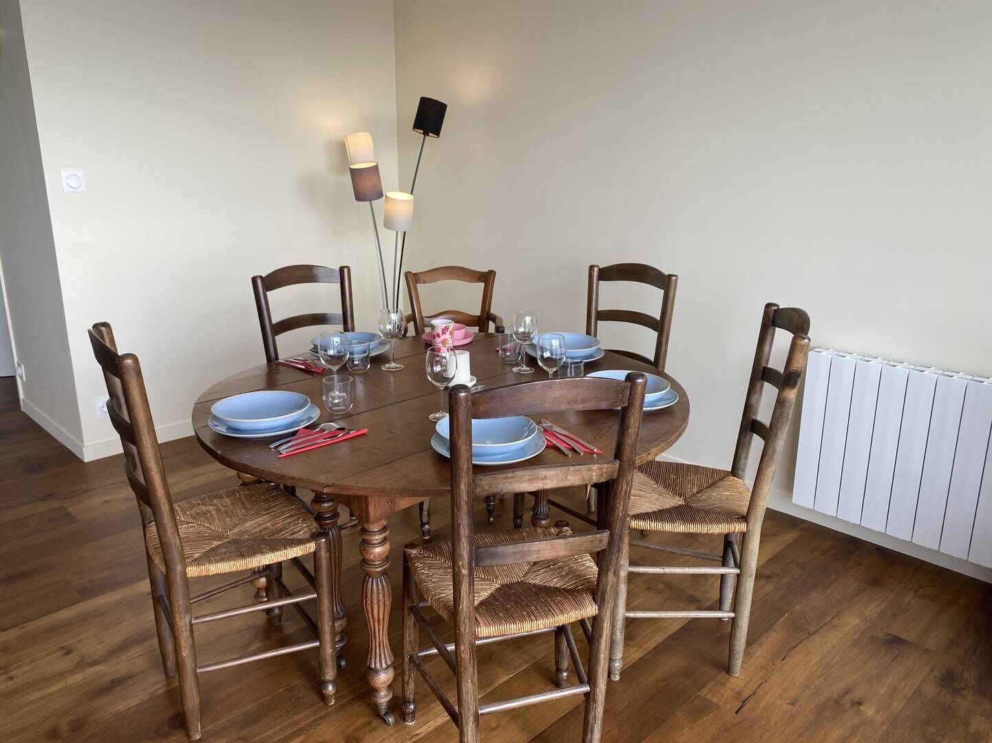 Une table à manger pour six personnes, avec des chaises en bois, un radiateur blanc et un luminaire fixé au mur.