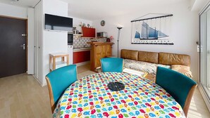 Un espace de vie compact comprenant un coin cuisine, un espace repas avec une table aux couleurs vives et un canapé recouvert d’un tissu à motifs.