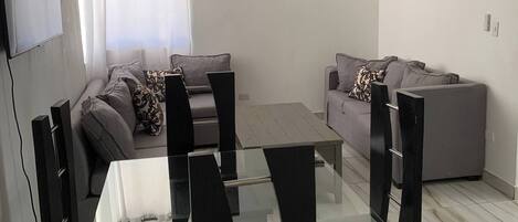 A modern living room with a glass dining table, black chairs, a grey sofa, and a TV mounted on the wall.