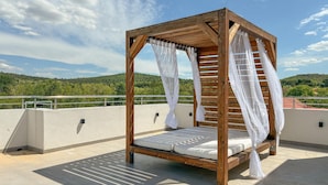 Uma cama de madeira com dossel e cortinas brancas em um terraço no telhado, com vista para colinas verdes.