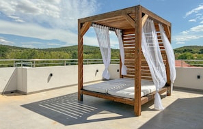Ein hölzernes Baldachinbett mit weißen Vorhängen auf einer Dachterrasse mit Blick auf grüne Hügel.