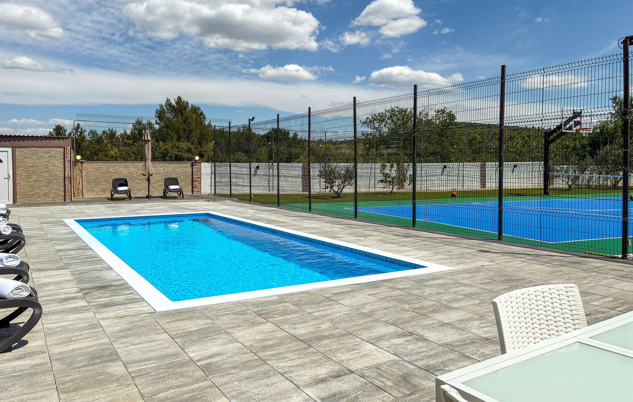 A swimming pool with a netted area, lounge chairs, and a tennis court.