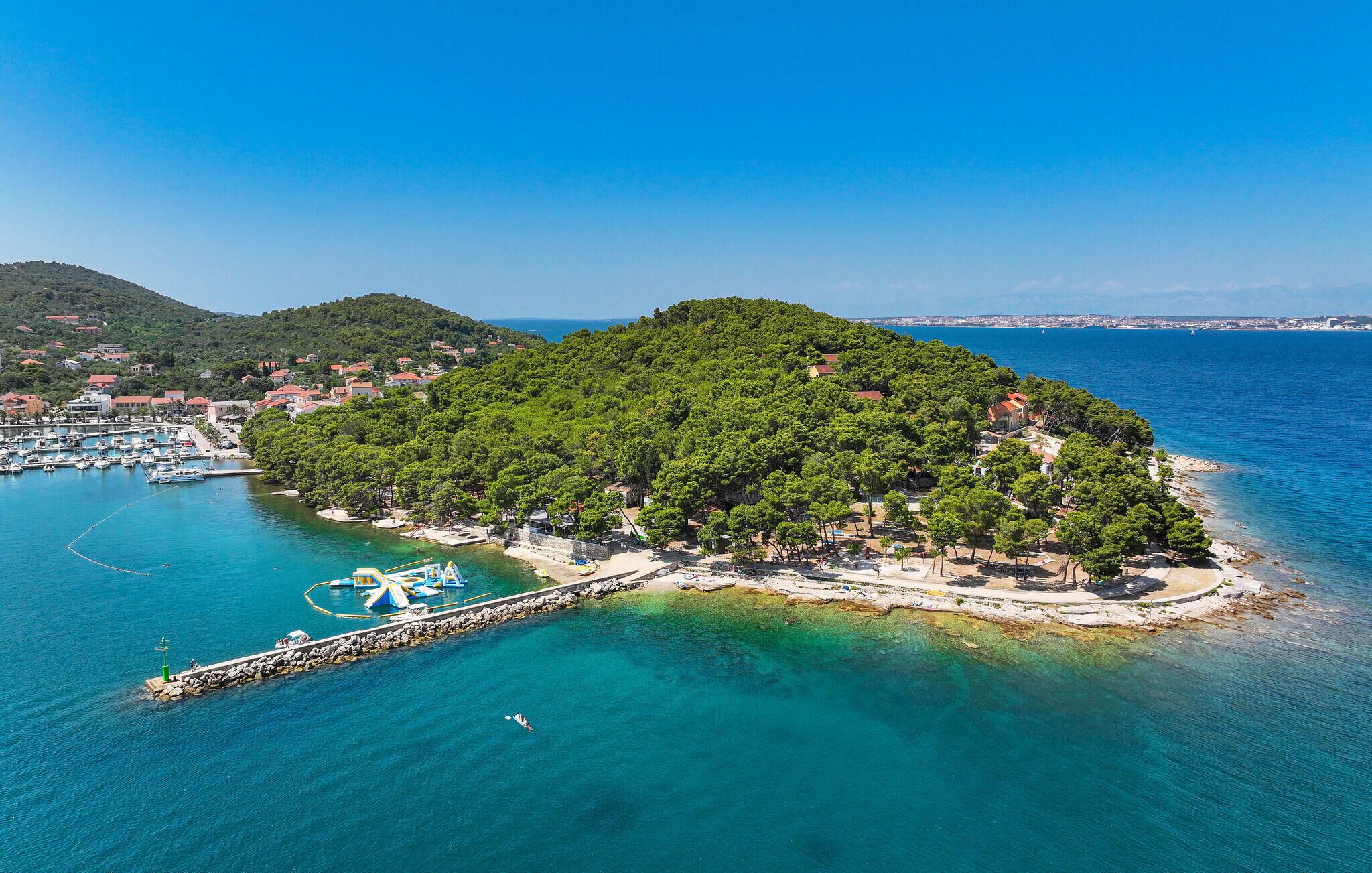 Une vue aérienne d’une petite île dotée d’un port de plaisance, entourée d’eaux d’un bleu limpide, avec une ville lointaine en arrière-plan.