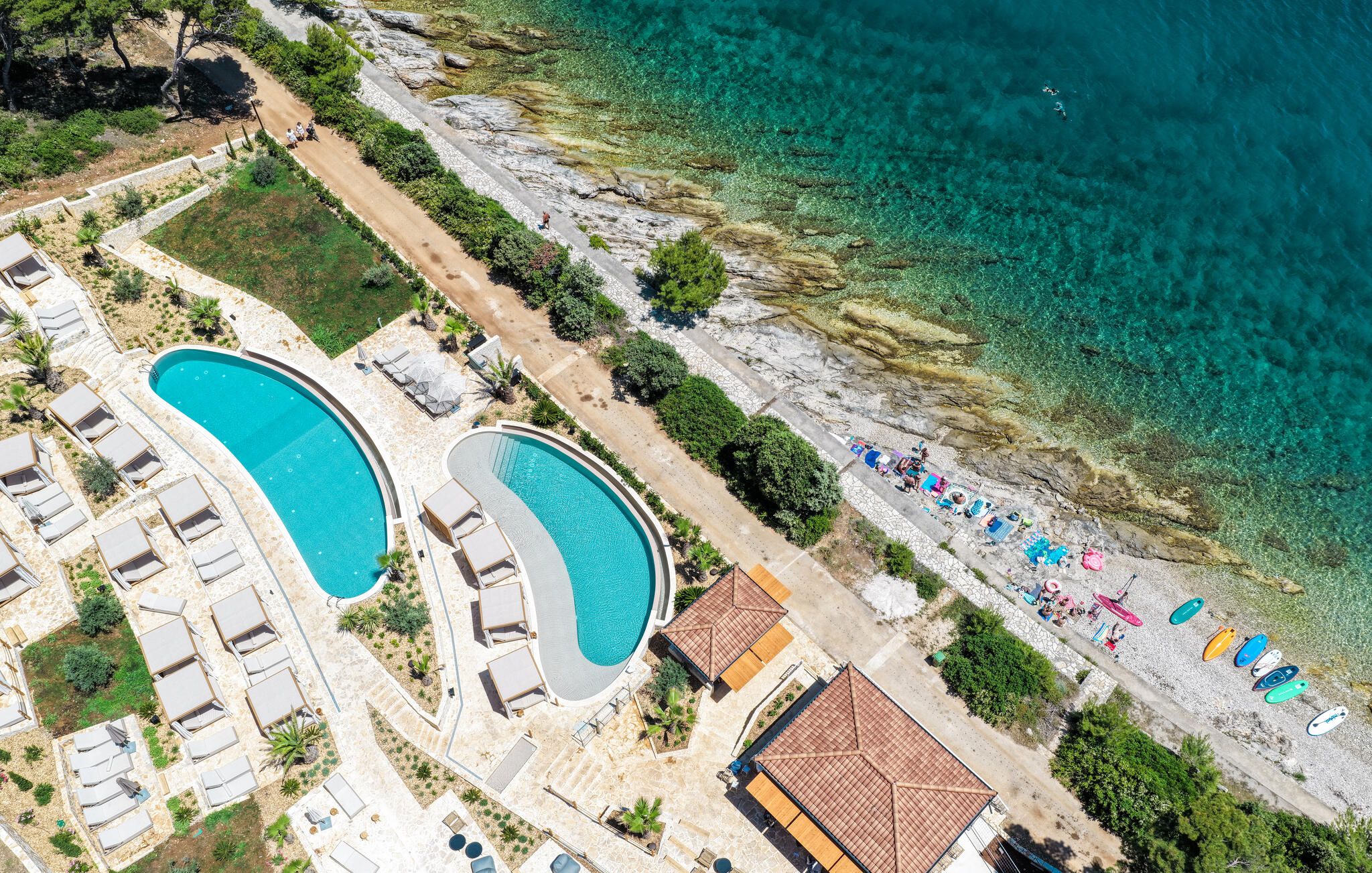 Eine Luftaufnahme eines Resorts mit zwei großen Schwimmbecken, einem Strandabschnitt mit Sonnenschirmen und einer felsigen Küstenlinie.