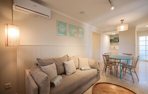 Ein Wohnzimmer mit einem beigefarbenen Sofa, einem Esstisch mit Stühlen und einer Küche im Hintergrund.