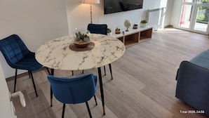 Ein modernes Wohnzimmer mit einem runden Marmortisch, blauen Stühlen, einem Fernseher und einer Glastreppe mit Pflanzen.