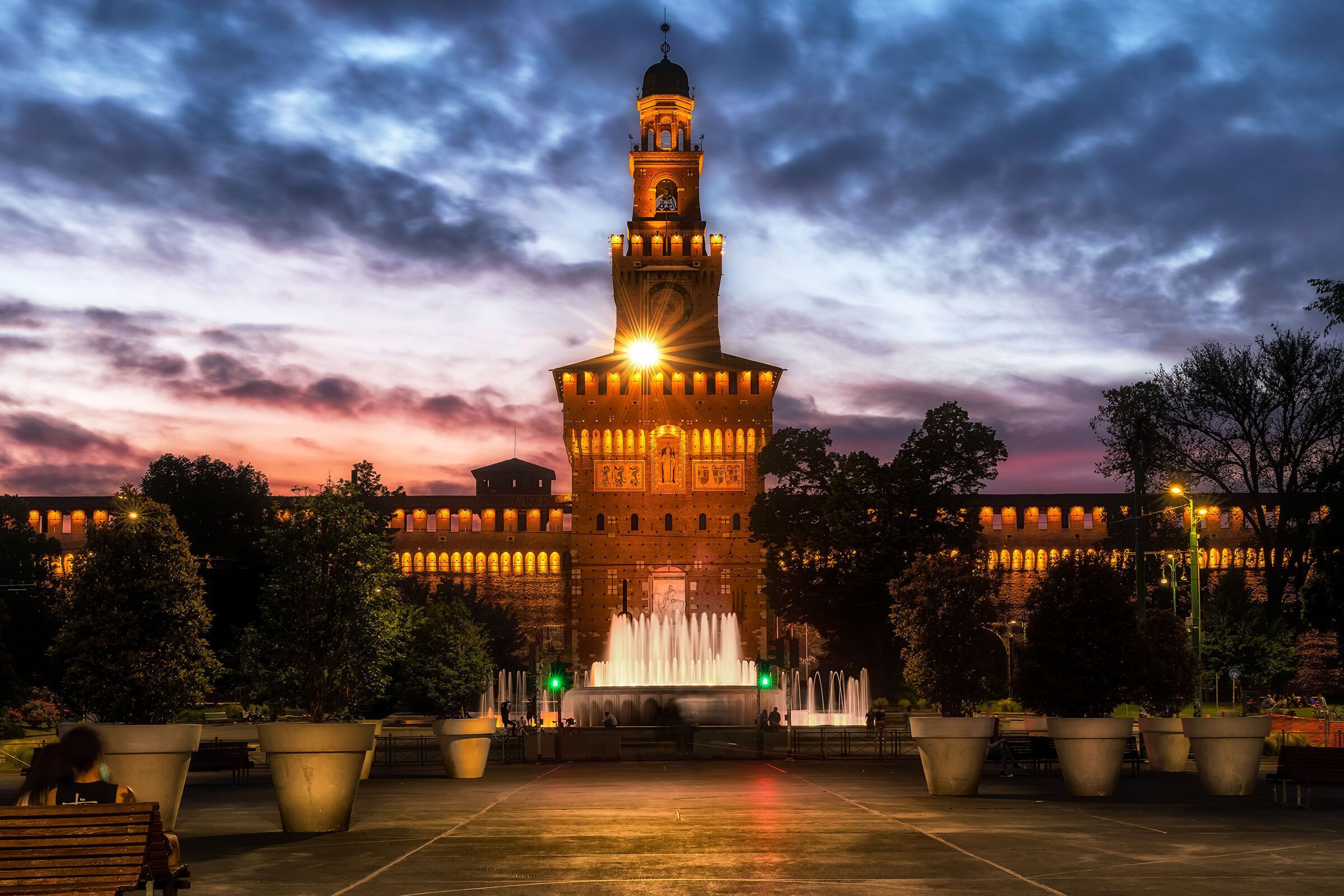 Una torre histórica con un reloj, rodeada por una plaza con macetas grandes y una fuente.