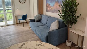 Ein modernes Wohnzimmer mit einem grauen Sofa, einem runden Tisch, einer Zimmerpflanze und einem Gemälde, das ein Gebäude zeigt.
