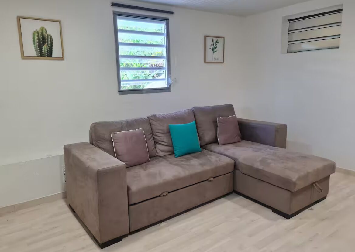 A beige sectional sofa with a teal pillow, a window with blinds, and two framed pictures on the wall.