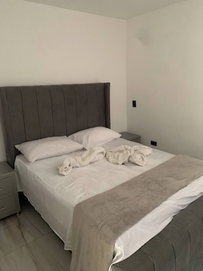 Cama bem arrumada com cabeceira cinzenta, almofadas brancas e manta bege.