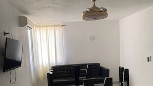 Ein modernes Esszimmer mit einem Glastisch und Stühlen, einem schwarzen Sofa, einem an der Wand befestigten Flachbildfernseher und einer Deckenleuchte.