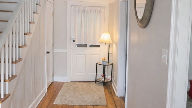Un pasillo con piso de madera, una escalera, una puerta con marco blanco, una lámpara sobre una mesita y un espejo colgado en la pared.