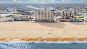 A coastal cityscape with a sandy beach, a large hotel with multiple floors, and a bridge in the background.