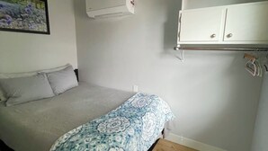 A bedroom with a bed, a ceiling fan, an air conditioning unit, a wardrobe, and a painting on the wall.