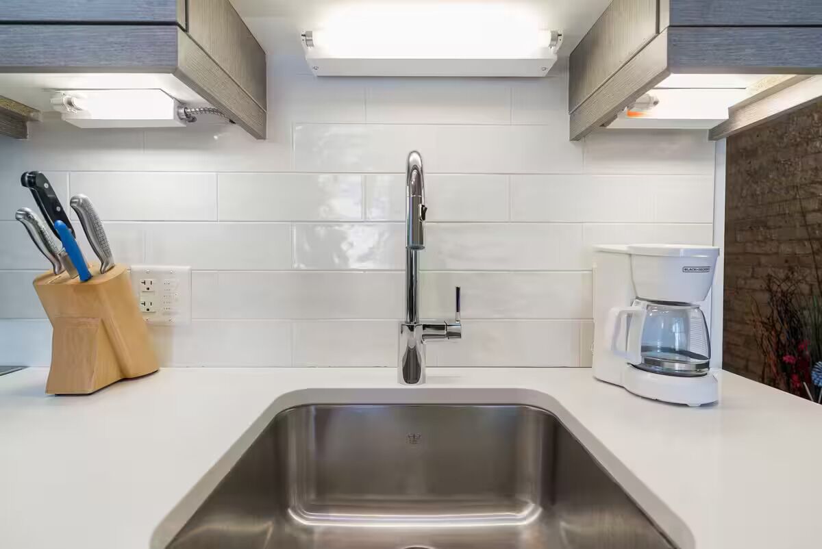 Una cocina moderna con un lavamanos de acero inoxidable, salpicadero de azulejos blancos y una cafetera sobre la encimera.