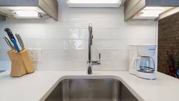 Una cocina moderna con un lavamanos de acero inoxidable, salpicadero de azulejos blancos y una cafetera sobre la encimera.