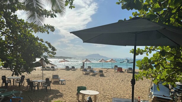 A beachside seating area with tables and chairs, umbrellas, and a view of the sea and distant mountains. - Condo Bien Situé 3 Minutes de la Plage a Pied 312 (Sosúa)