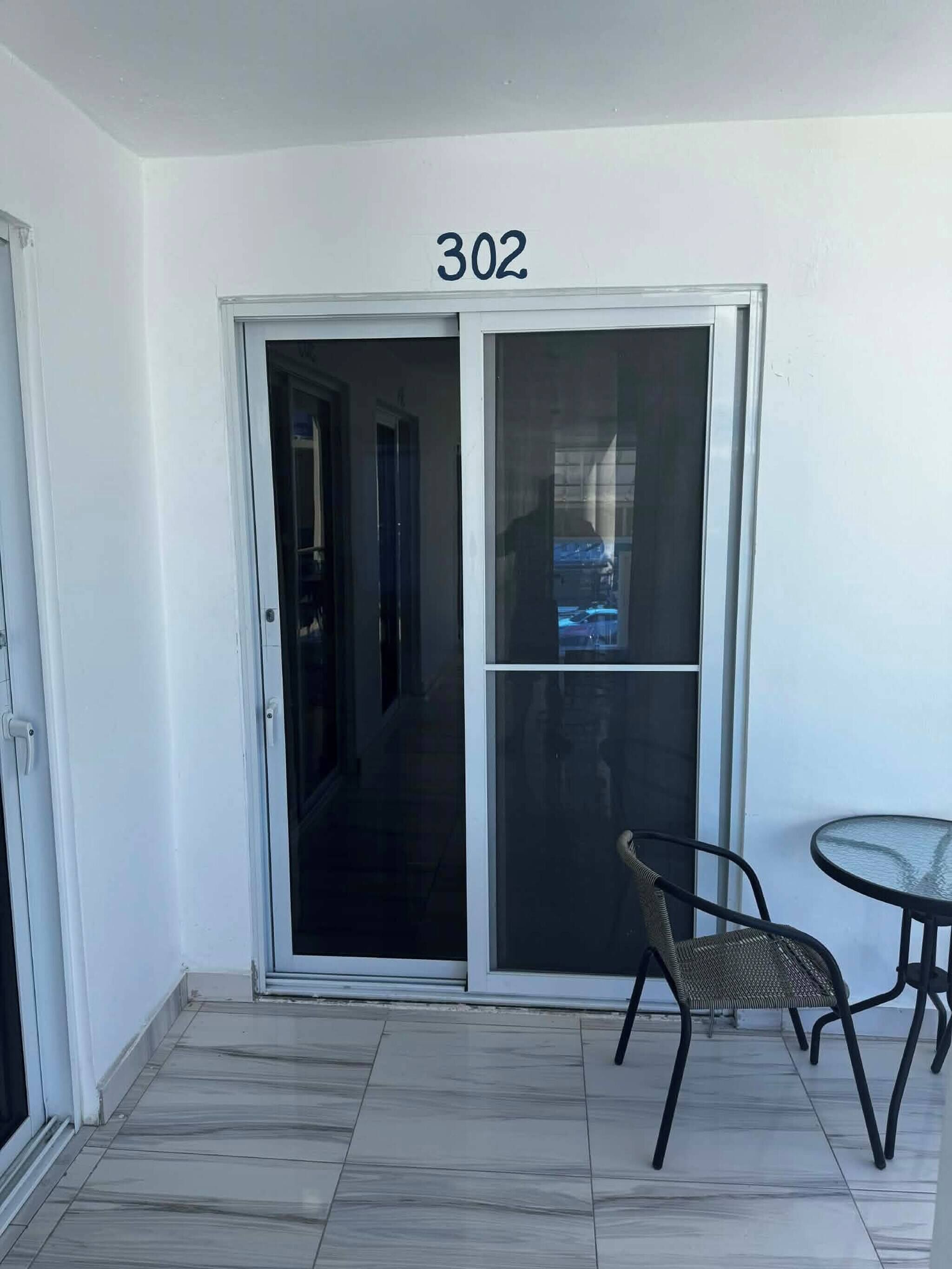 A room with a sliding glass door marked '302,' a small round table, and a wicker chair.