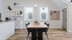 Een moderne keuken met witte kasten, een houten eettafel en zwarte stoelen. Er zijn twee ramen met zonwering, een hanglamp en een schilderij aan de muur.