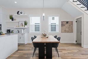 A modern kitchen with white cabinetry, a wooden dining table, and black chairs. There are two windows with blinds, a pendant light, and a painting on the wall. - Cozy, Modern Retreat in the Heart of College Park Perfect for Families & Guests! (College Park)