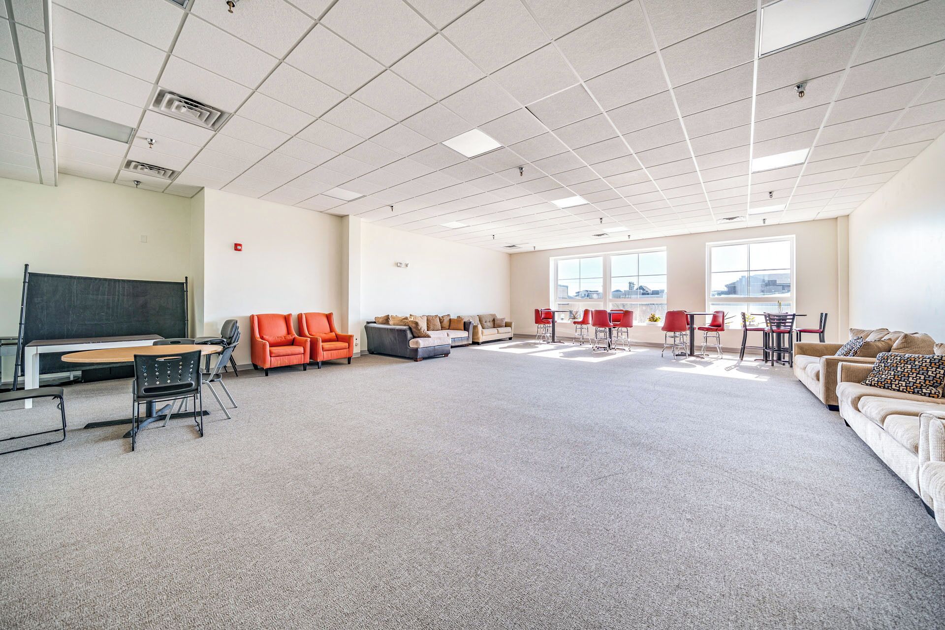 A spacious room with a large carpeted floor, a blackboard, orange chairs, a sofa, and a dining area with red chairs.