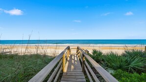 Un sentier en bois mène à une plage où l’eau est d’un bleu cristallin et le rivage est sablonneux.