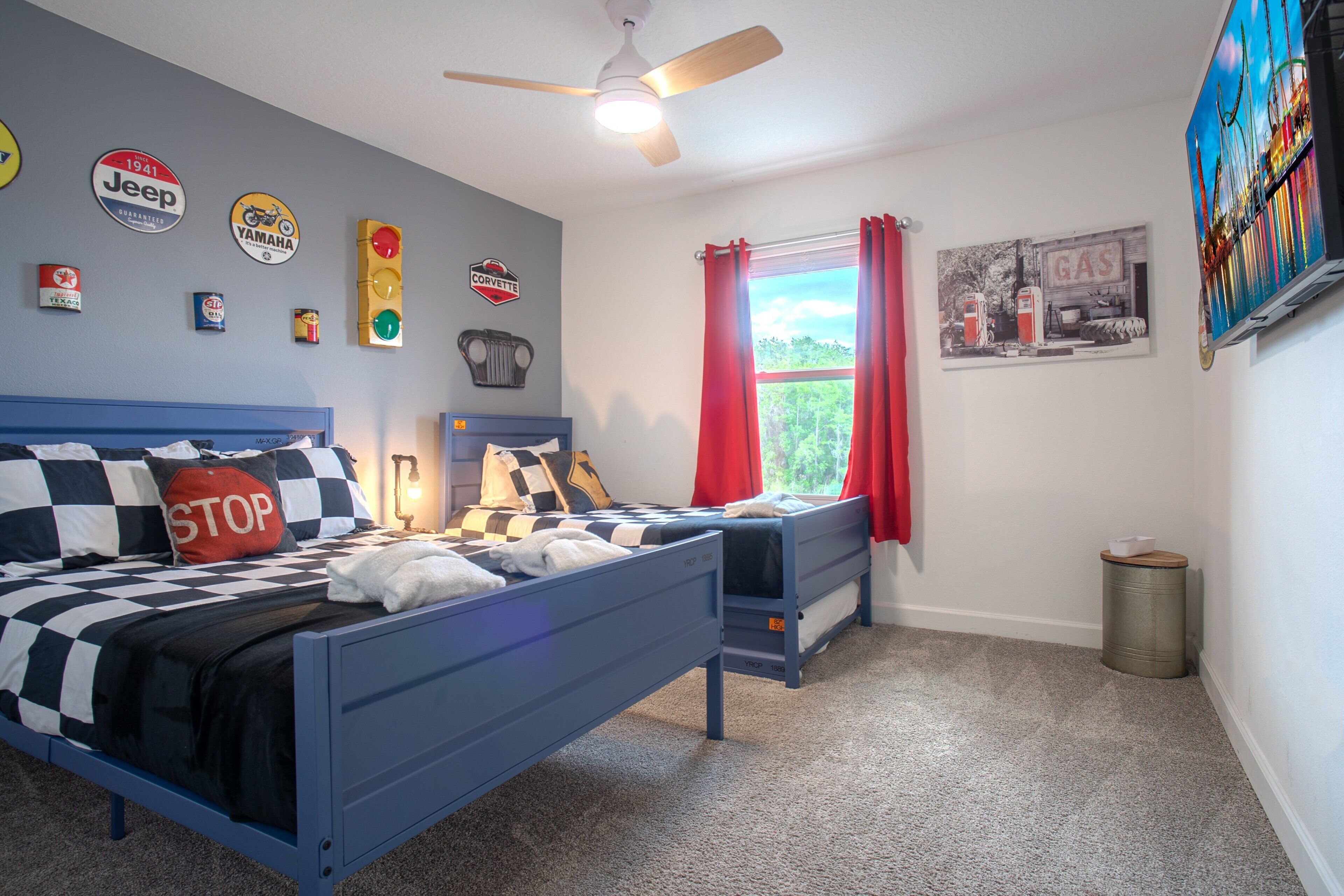 A bedroom with a bed, a smaller bed, a ceiling fan, a window with red curtains, and wall decorations including vintage car signs.