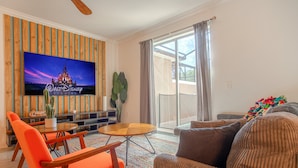 A living room with a flat-screen TV, orange chairs, a brown sofa, and a large window. - Spacious 4BR Townhome w/ Private Pool • Storey Lake Resort • Near Disney (Kissimmee)