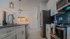 A modern kitchen with stainless steel appliances, a central island, pendant lighting, and a view into the dining area. - Spacious 4BR Townhome w/ Private Pool • Storey Lake Resort • Near Disney (Kissimmee)