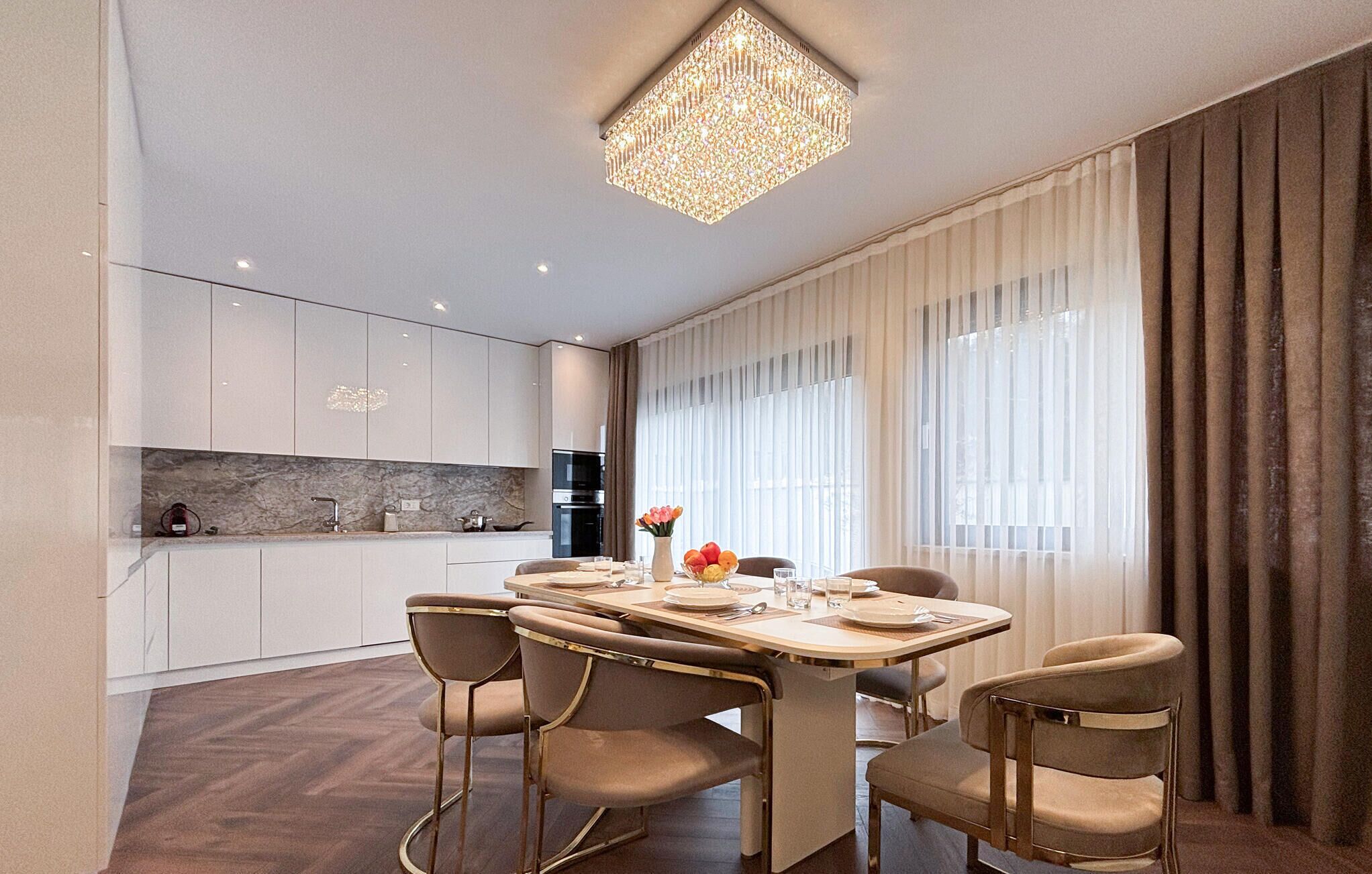 A modern dining area with a chandelier, a dining table set for four, and a kitchen in the background.