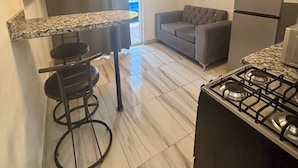A compact kitchen with a gas stove, a bar stool, a grey sofa, and a ceiling light fixture. - Condo Neuf Dans le Centre-ville de Sosua 104 (Sosúa)