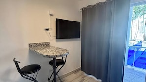 A room with a marble-tiled floor, a round ceiling light, a flat-screen TV mounted on the wall, a bar area with a granite countertop and two black bar stools, and a large window with a view of a blue pool and greenery. - Condo Neuf Dans le Centre-ville de Sosua 104 (Sosúa)