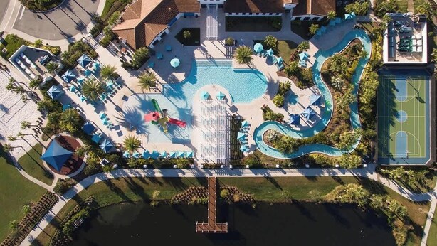Eine Vogelperspektive zeigt ein Resort mit einem zentralen Schwimmbecken, einem Tennisplatz und einer Anlegestelle am Wasser.