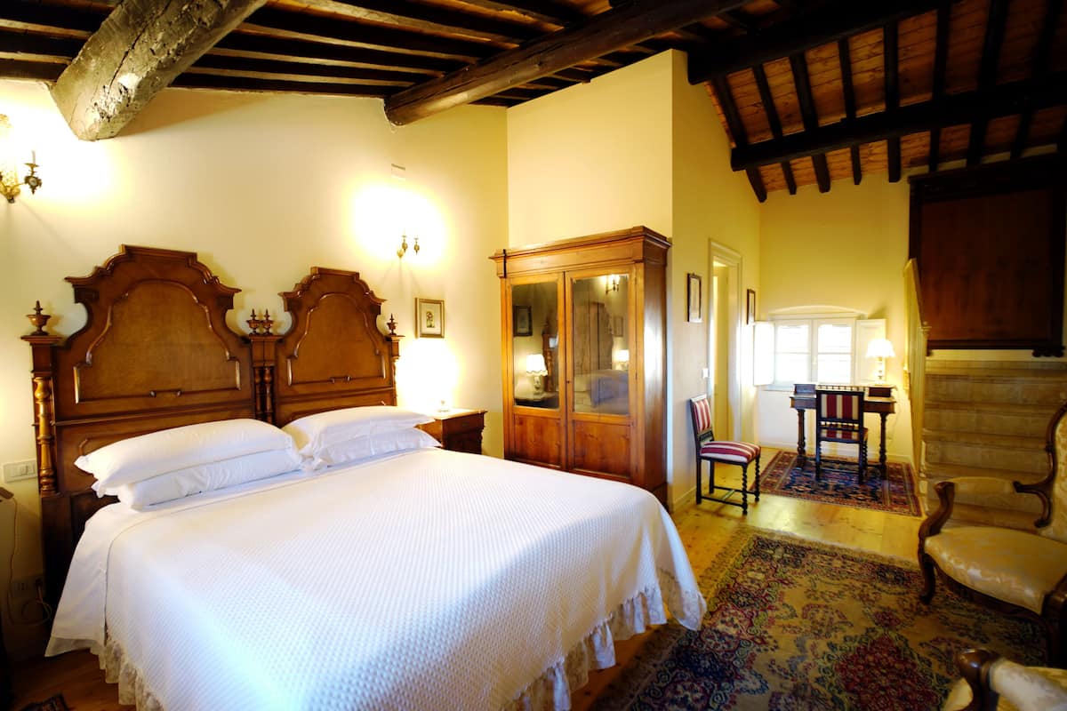 A bedroom with a large bed, wooden headboard, a wooden wardrobe, a chair, and a table with a lamp.