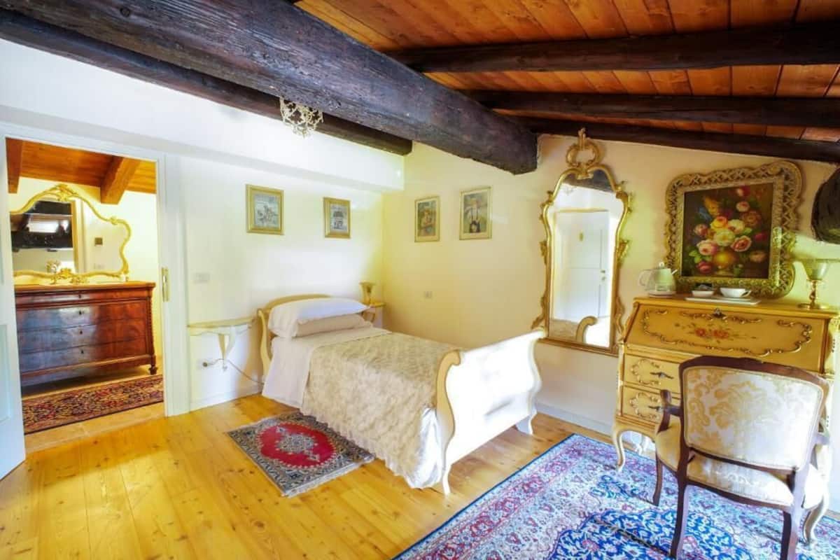A bedroom with a wooden ceiling, a bed with a white bedspread, a wooden dresser, a chair, and a mirror with a floral painting.