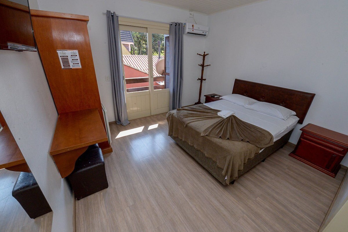 A hotel room with a bed, a wooden wardrobe, a small bedside table, a heater, and a view of the outside.