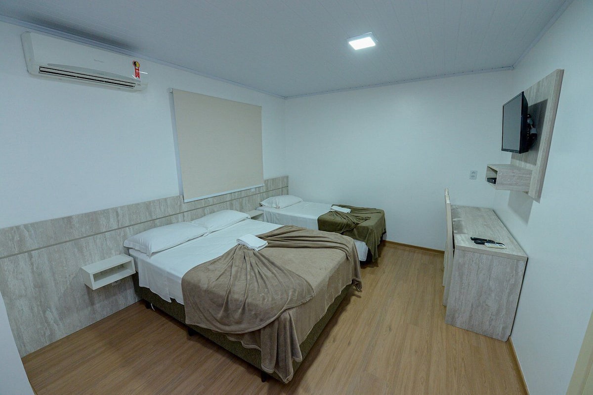 A hotel room with two beds, a small desk, a TV mounted on the wall, and a window with blinds.