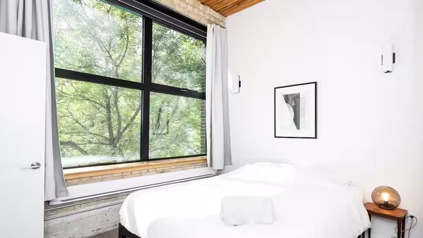 Un dormitorio con un ventanal grande, una cama, una mesita de noche con lámpara y un cuadro enmarcado en la pared.