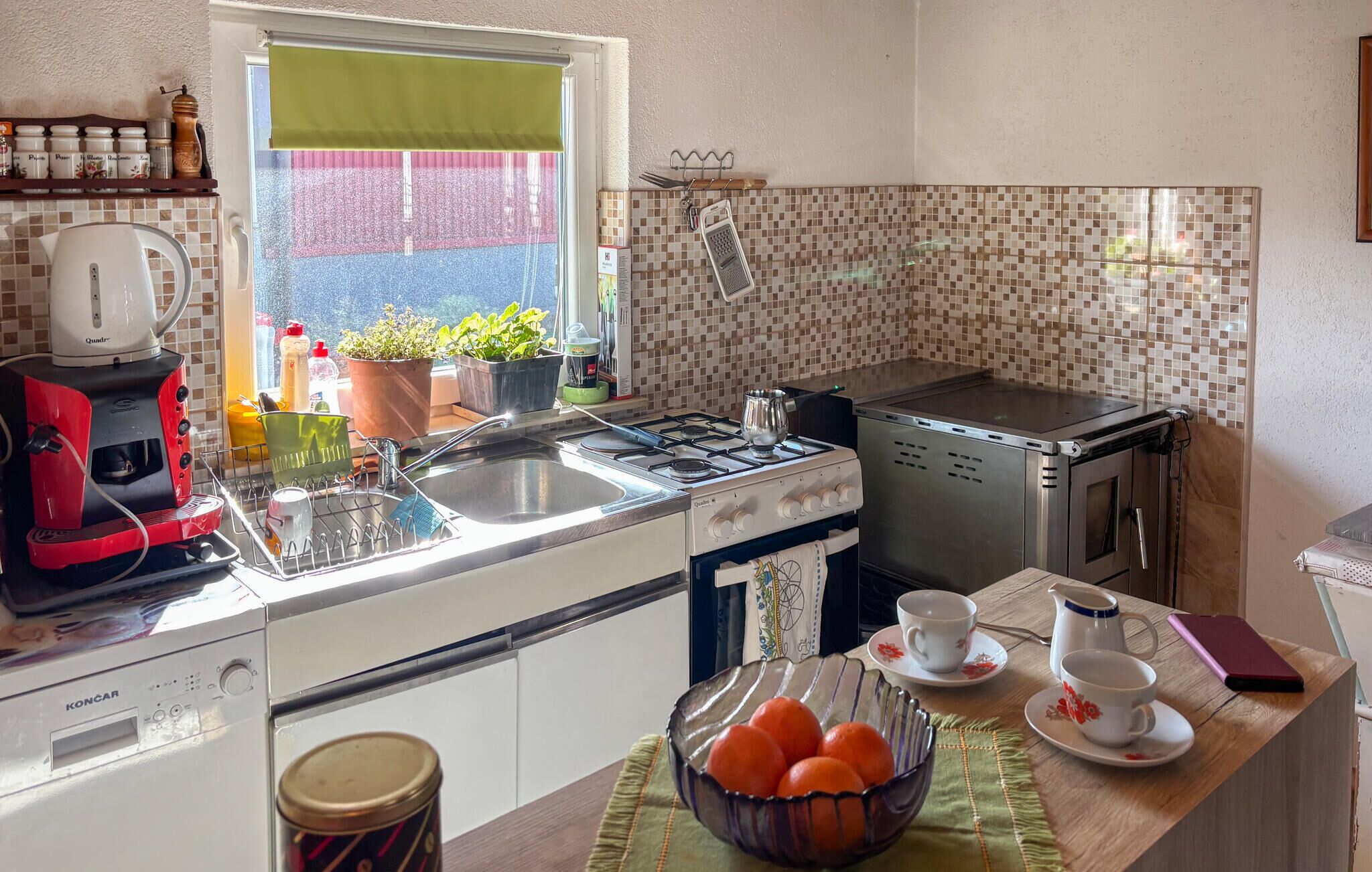 Una cocina con estufa a gas, hervidor y cafetera. Hay plantas en macetas en el alféizar de la ventana y un cuenco con naranjas en la encimera.