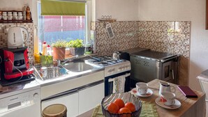 Eine Küche mit Gasherd, Wasserkocher und Kaffeemaschine. Auf dem Fensterbrett stehen Blumentöpfe und auf der Arbeitsfläche liegt eine Schüssel mit Orangen.