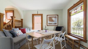 Una sala de estar acogedora con una escalera de madera, una mesa de comedor con sillas, un sofá con cojines y un amplio ventanal con vistas al exterior.