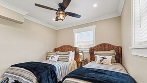 Une chambre avec deux lits, un ventilateur de plafond, une fenêtre avec des stores et une table de chevet avec une lampe.