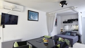 A compact hotel room with a dining table, two chairs, a bed, a TV, and a ceiling fan. - ENSO - Studio Héliopolis J 4 + garage - Luxe - 2 people (AGDE)
