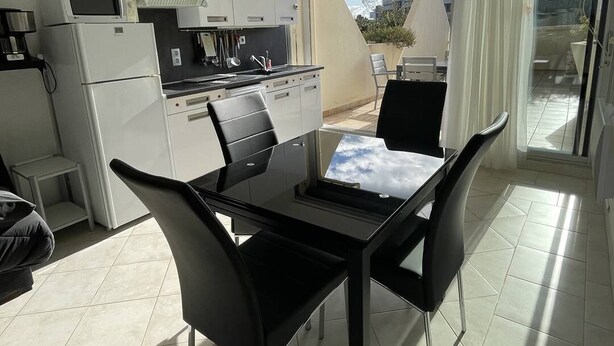 A modern kitchen with white cabinets, a black countertop, and a dining area with a glass table and black chairs. - ENSO - Studio Héliopolis J 4 + garage - Luxe - 2 people (AGDE)
