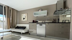 A modern kitchen with a built-in dishwasher, a flat-screen TV, and a dining area with a glass table and a sofa. - KERYLOS - Studio Héliopolis K 3 with garage - Luxe - 2 people (AGDE)